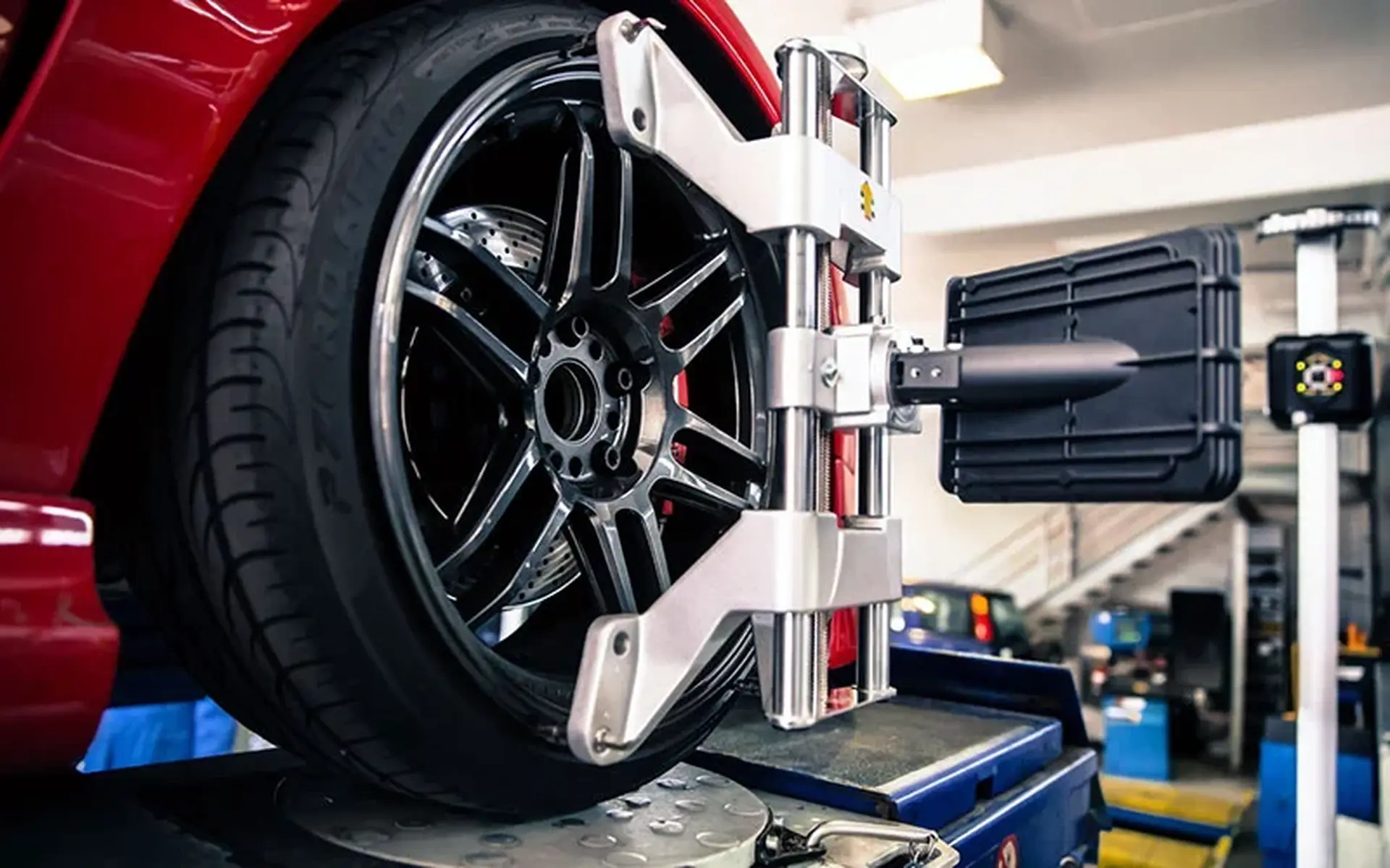 Car wheel on an alignment machine in a garage, with a red car and a digital alignment tool attached to the wheel.