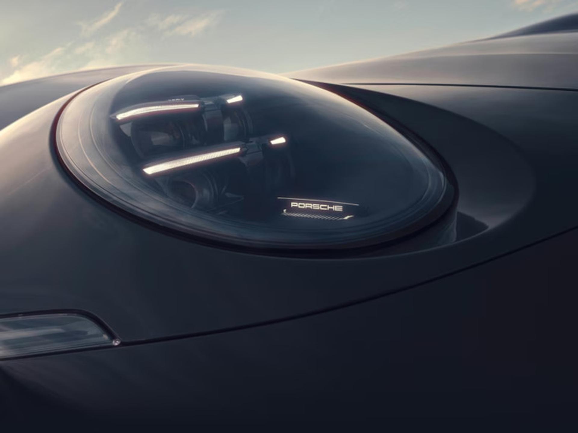Close-up of a sleek car headlight with the Porsche logo, featuring streamlined design and reflecting ambient light against a backdrop of sky.