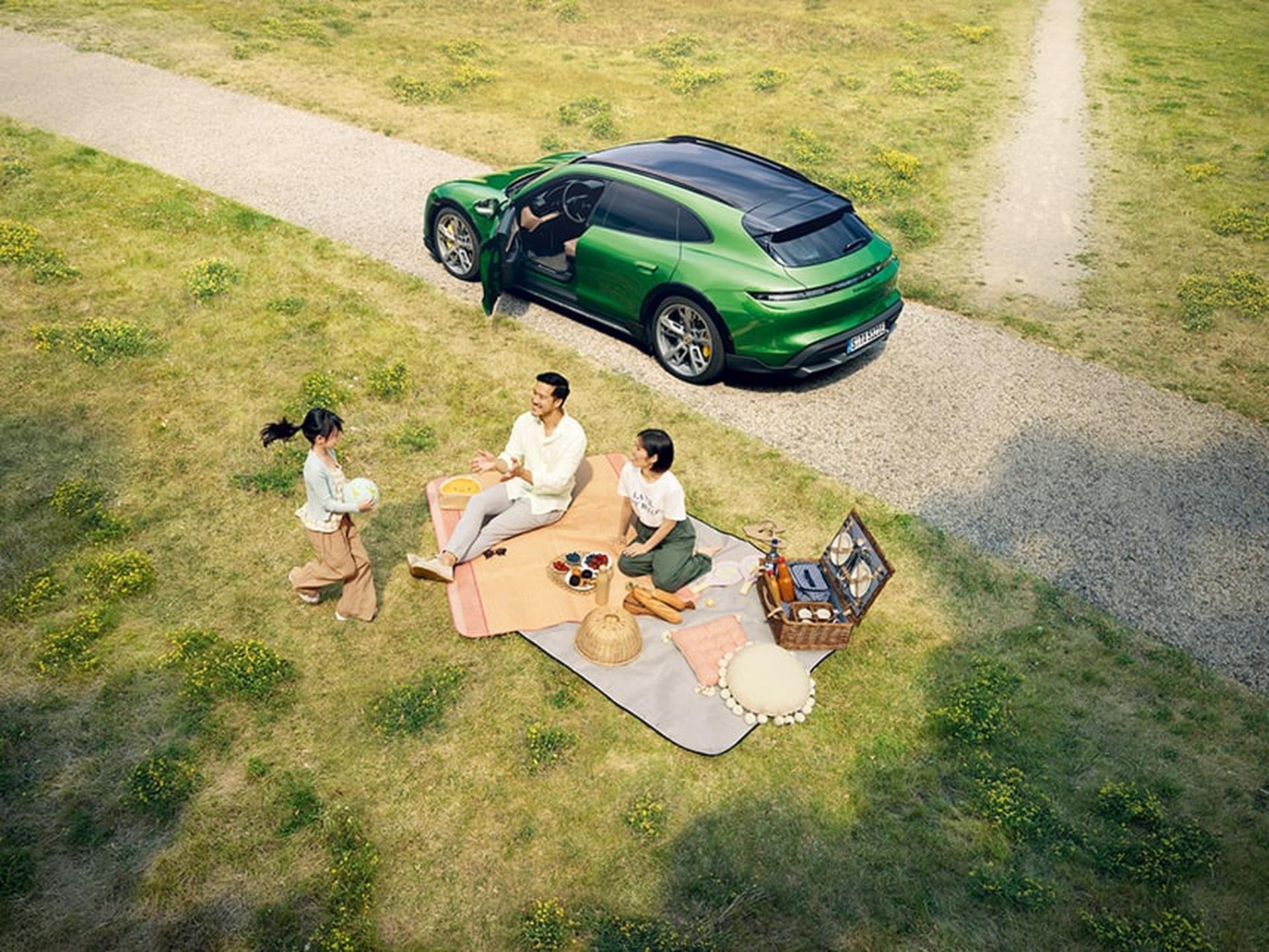 Eine Familie picknickt auf einer Wiese neben ihrem Porsche Sportwagen.