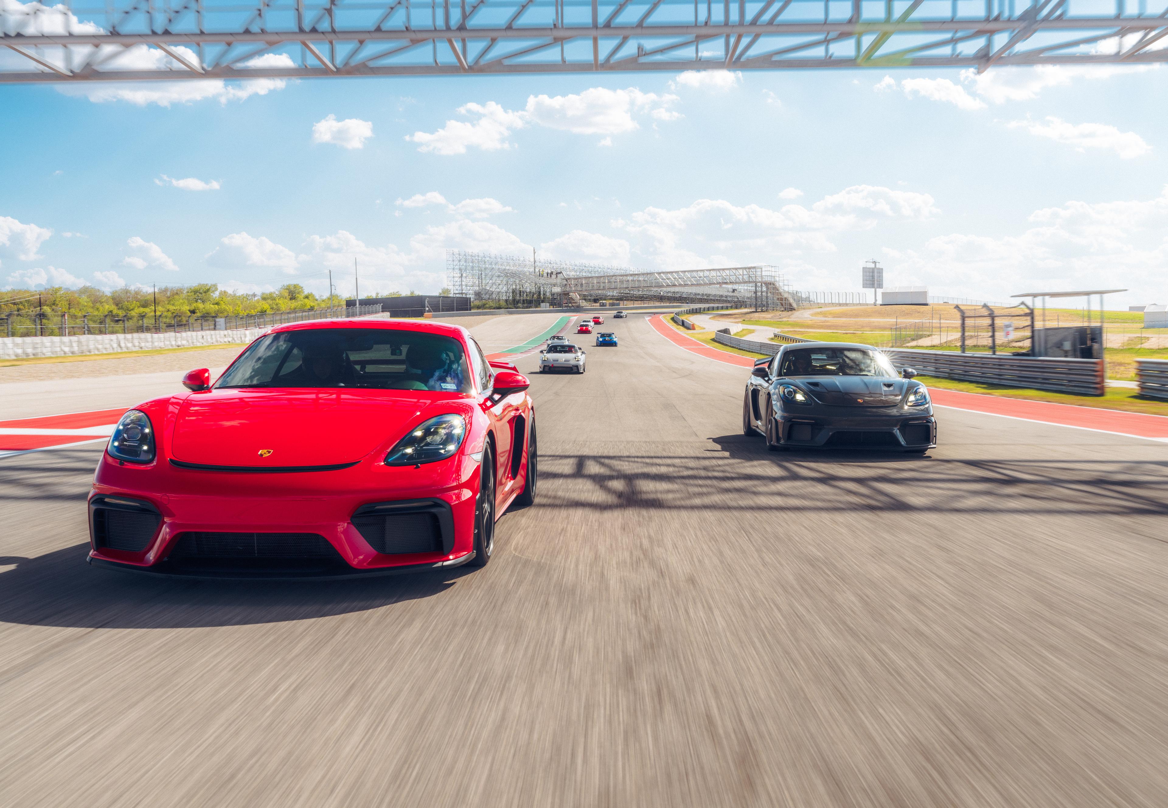 Red and black sports cars drive side by side on a sunny racetrack, with more cars trailing behind.