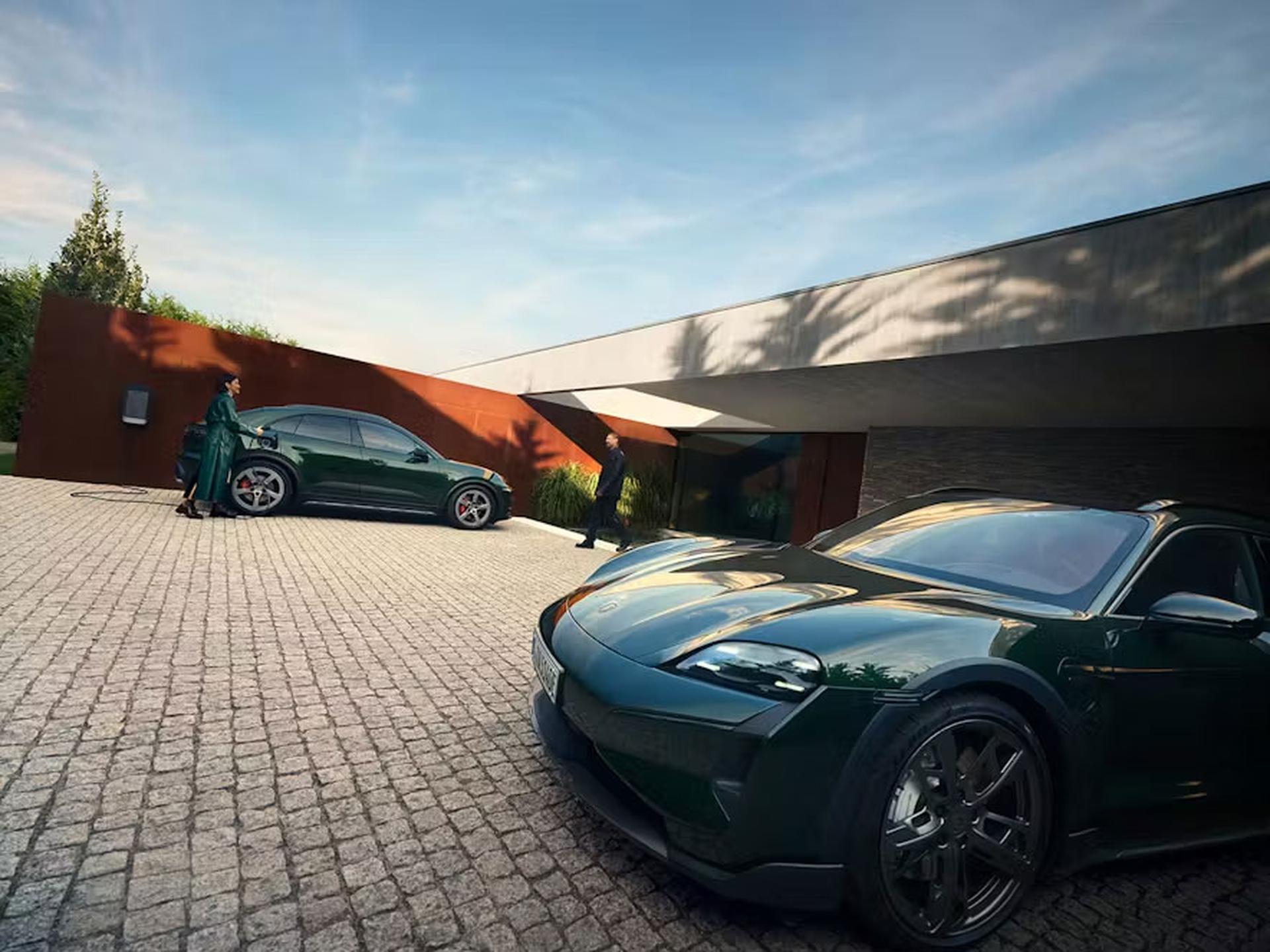 Zwei grün Porsche Fahrzeuge stehen auf einer modernen Auffahrt. Eine Frau lädt eines der Autos auf, während ein Mann unter klarem Himmel auf ein elegantes Haus zugeht.