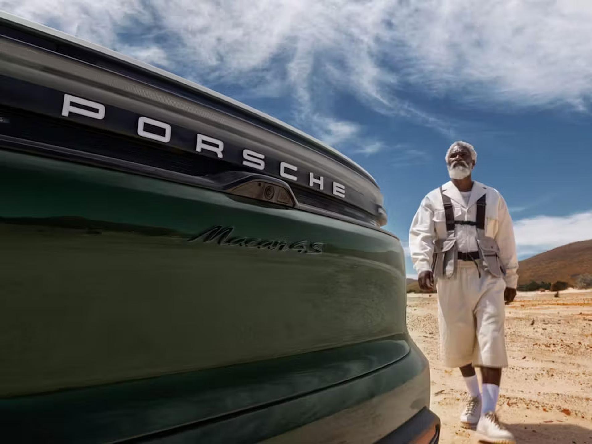 A man in a white outfit walks near a green Porsche Macan S in a desert landscape under a blue sky with clouds. Ein Mann in weißer Kleidung geht in einer Wüstenlandschaft unter blauem, bewölktem Himmel in der Nähe eines grünen Porsche Macan S.