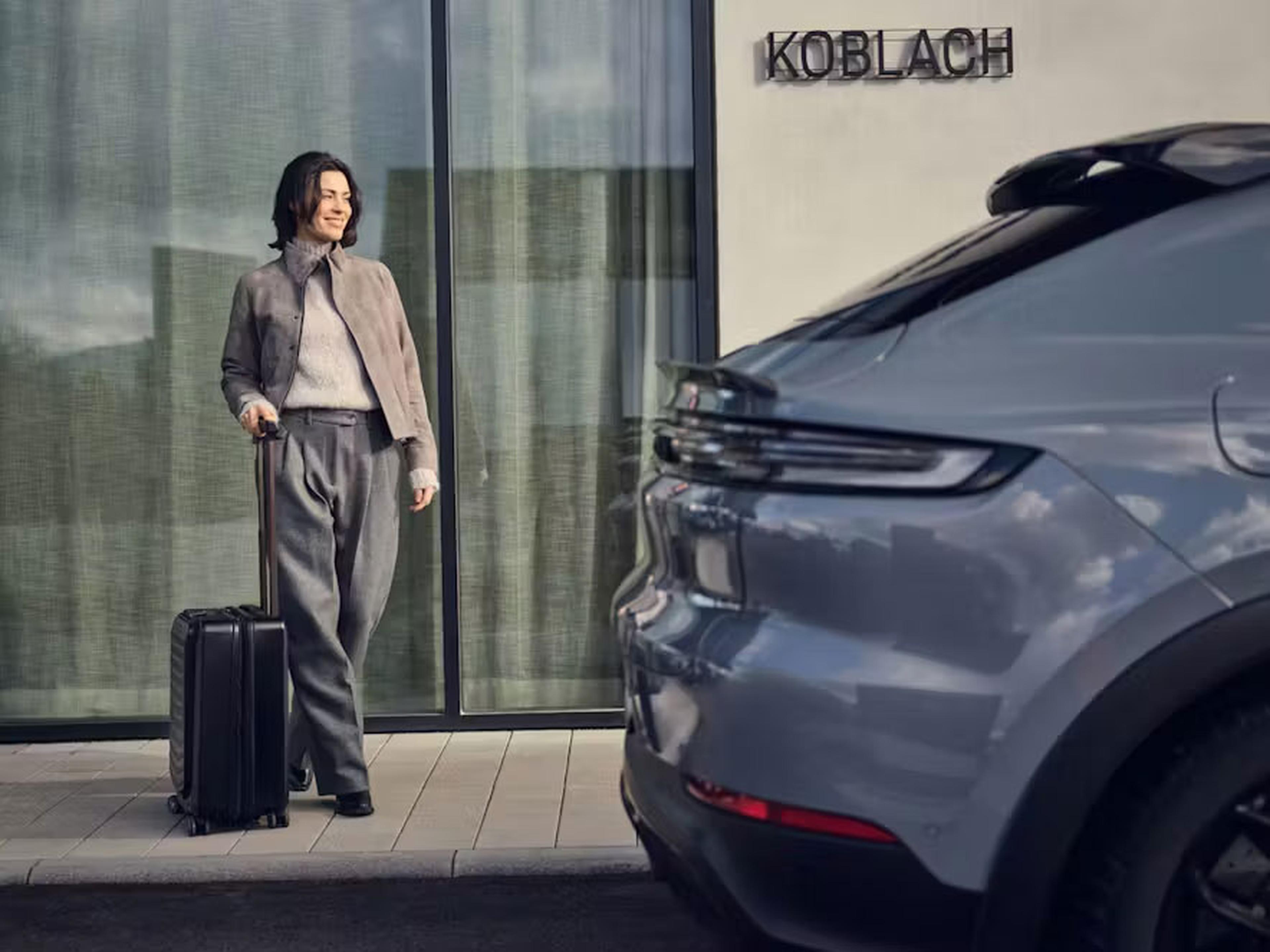 Eine grau gekleidete Person mit einem Koffer steht vor einem Gebäude, an dessen Wand „Koblach“ steht, und blickt auf ein geparktes Auto.