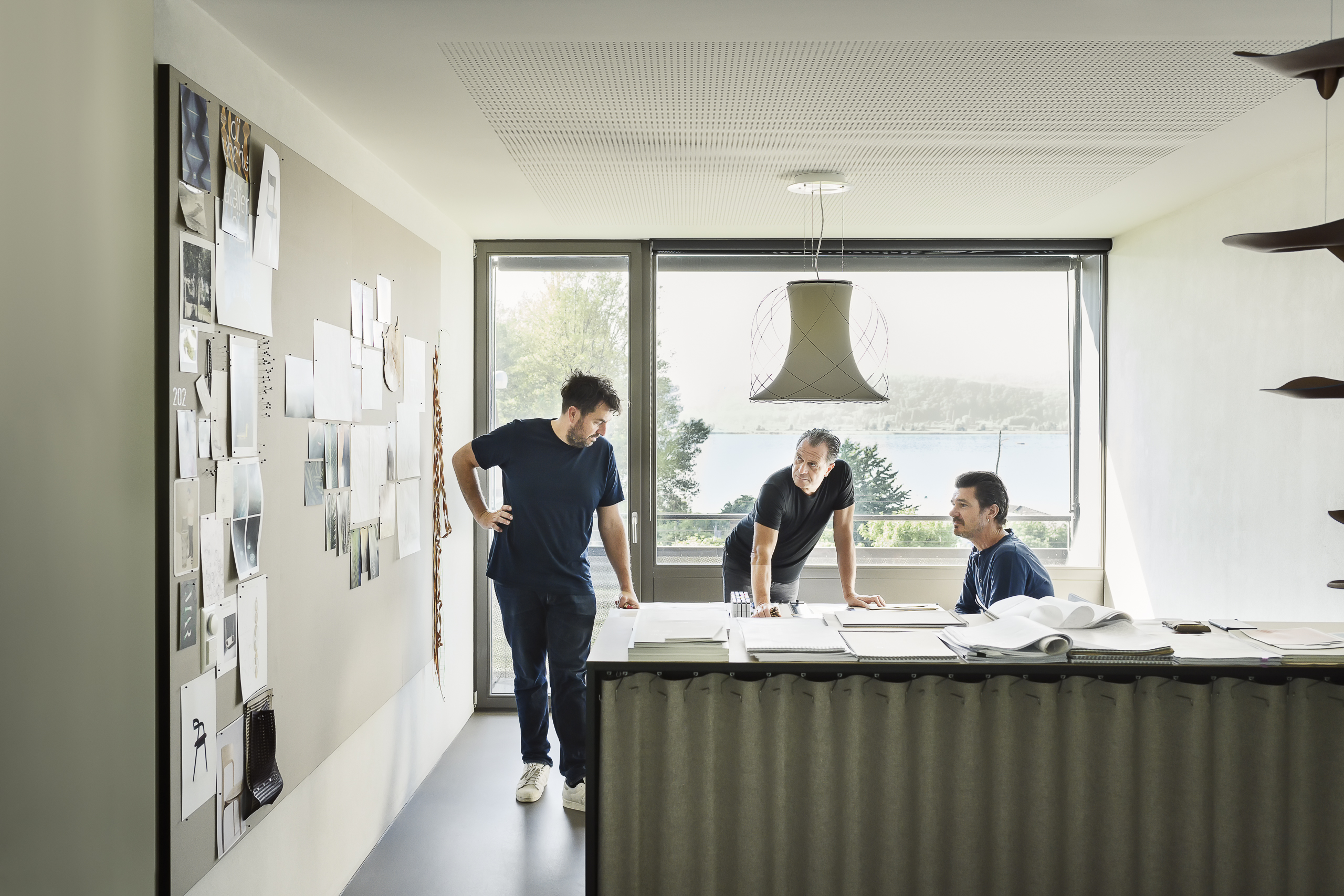 Three people in a bright design studio, discussing around a worktable in front of large windows.