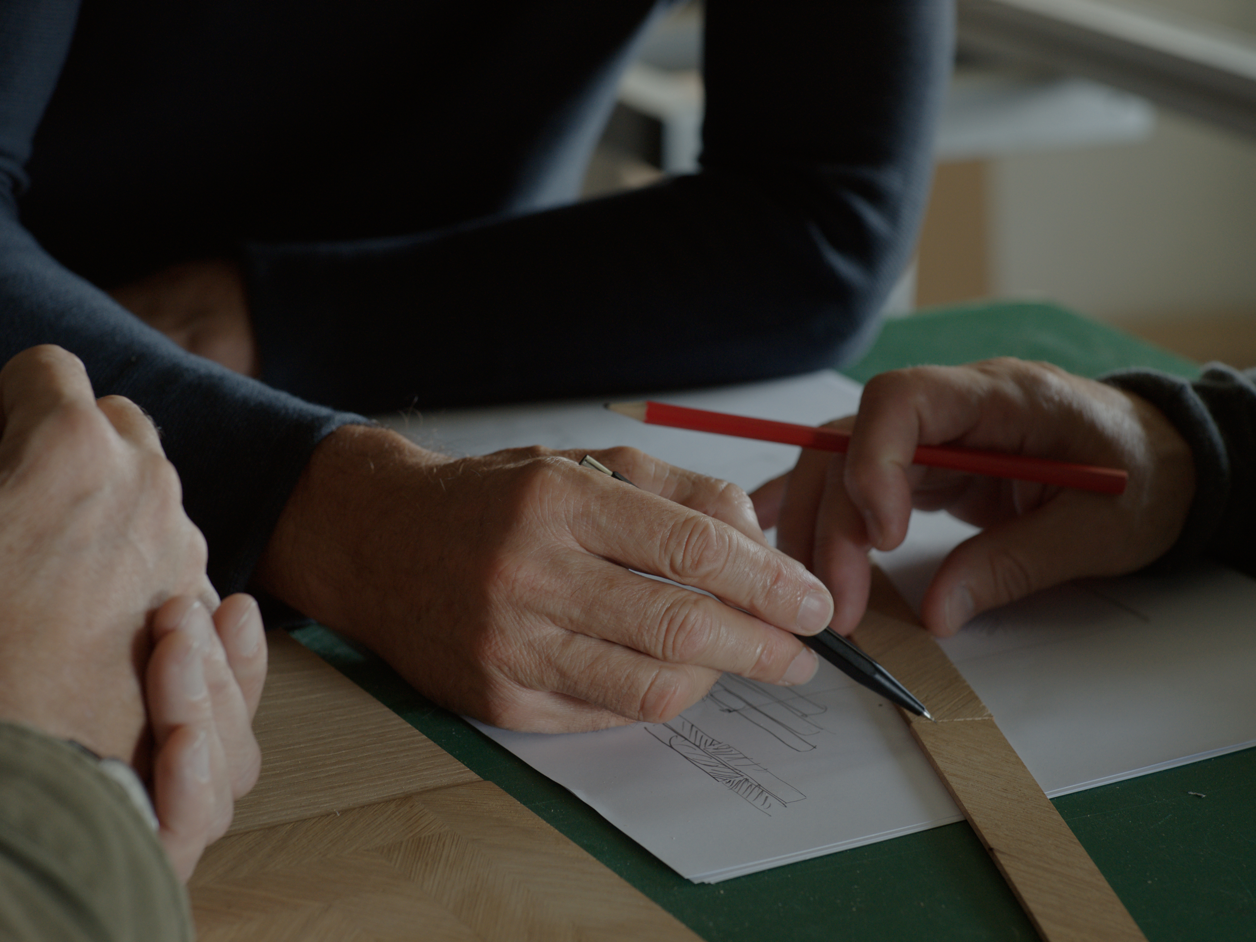 Hands sketching a parquet pattern on paper, using a wooden sample strip as a guide.