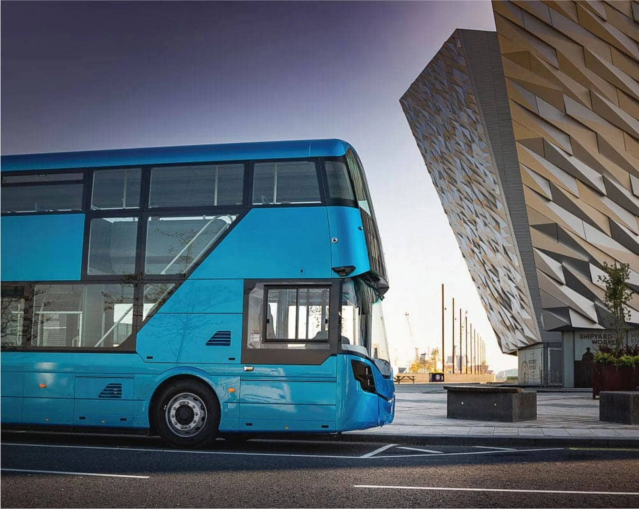 "Blue double-decker bus parked beside modern architectural building with textured facade in urban setting" [G]