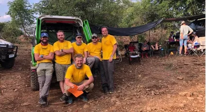 "Team in yellow shirts gathered at outdoor worksite with utility vehicle, under shade canopy in rural setting" [G]