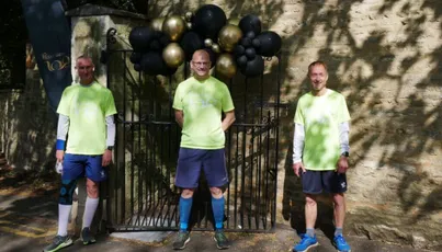 "Three team members in bright green shirts standing in front of a decorative gate with black and gold balloons for a Scott Bader event" [G]
