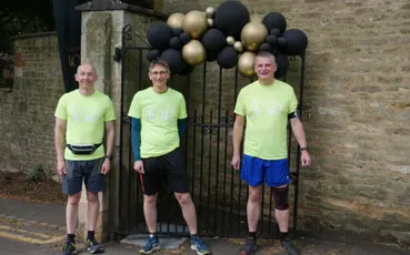 "Three people in bright yellow shirts standing by stone wall entrance with black and gold balloons for Scott Bader event" [G]