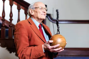 "Senior gentleman in orange blazer holding decorative sculpture award with wooden base, representing Scott Bader's industry recognition" [G]