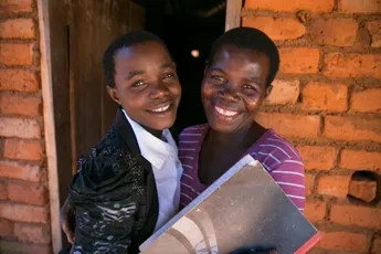 Two smiling students with notebooks standing in a brick doorway, representing education in a developing community. [G]