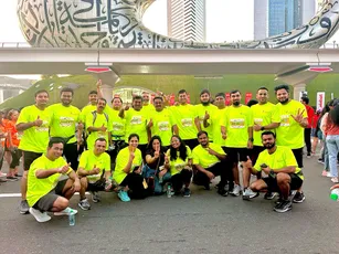 "Group of runners in bright yellow shirts gathered at an event in Dubai, with modern architecture and Arabic calligraphy visible in background." [G]