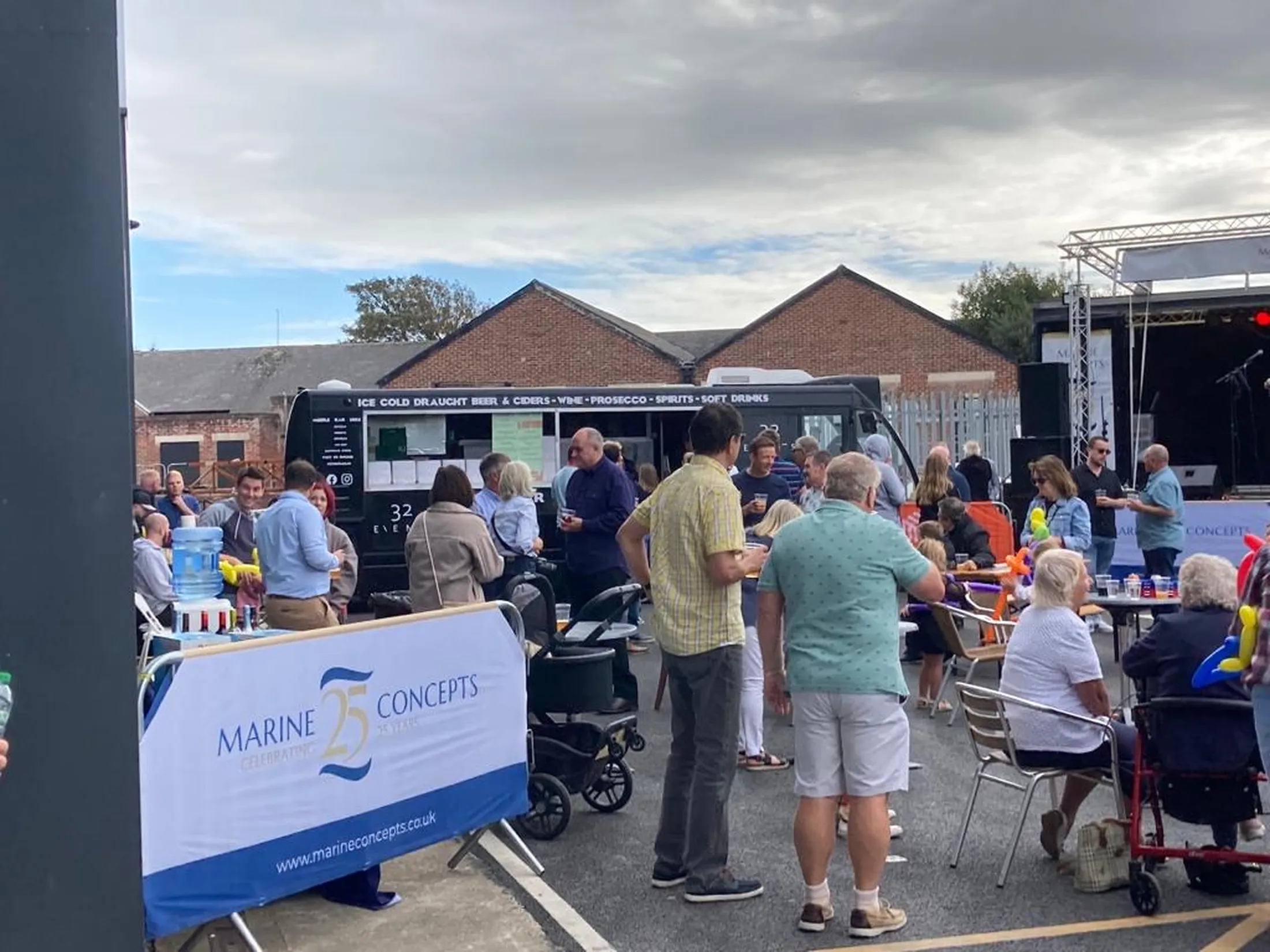 "Outdoor event with Marine Concepts banner and food truck, attendees gathered at tables under cloudy sky" [G]
