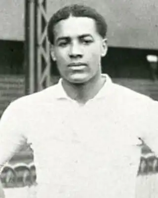 Historic black and white photograph of a person in a white shirt standing in front of stadium seating. [G]