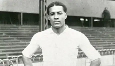 Historic black and white photograph of a person in a white shirt standing in front of stadium seating. [G]