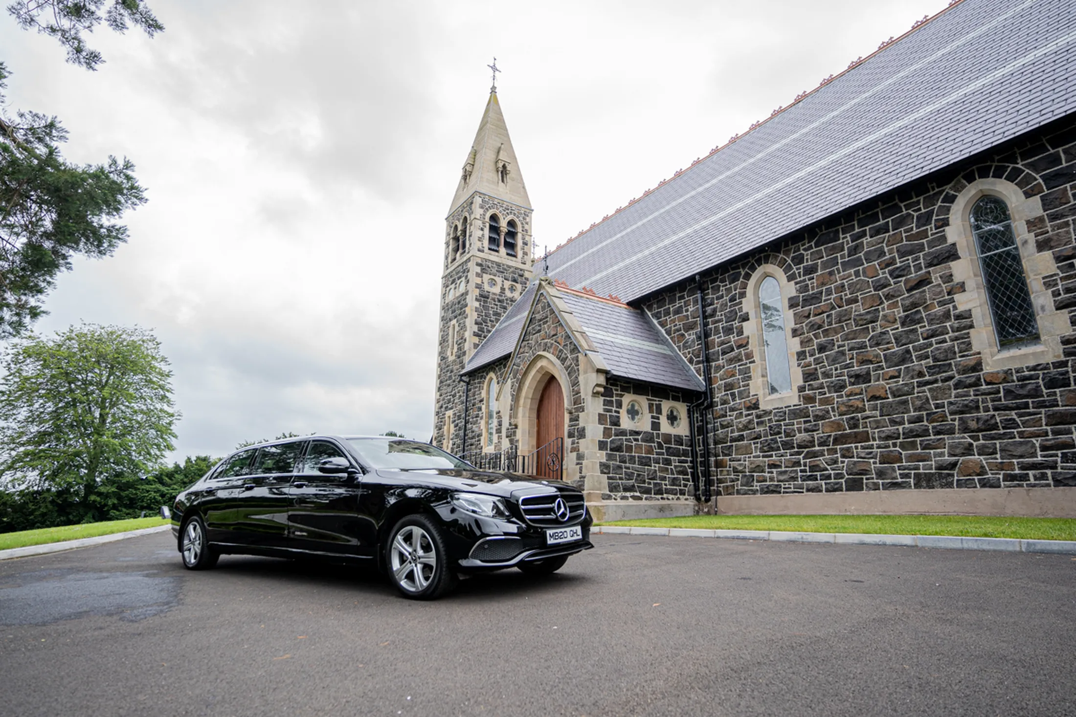 Black luxury car parked in front of a stone church with solar panels on the roof under cloudy sky. [G]
