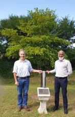 Two colleagues standing beside a commemorative plaque at Scott Bader, with green trees in the background. [G]