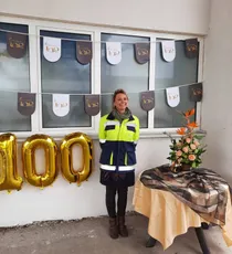 "Celebration event with decorative banners and gold '100' balloons at Scott Bader facility, featuring floral arrangement on table" [G]