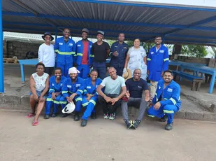 "Group of industrial workers in blue uniforms with safety reflective strips gathered under a metal shelter structure." [G]