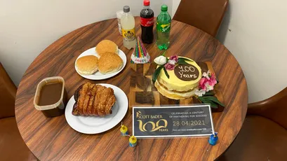 "Scott Bader 100th anniversary celebration with cake, pastries and drinks on wooden table featuring company logo" [G]