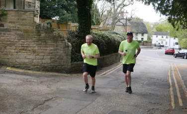 "Two runners in bright green shirts jogging along a village street with stone walls and cottages in the background." [G]