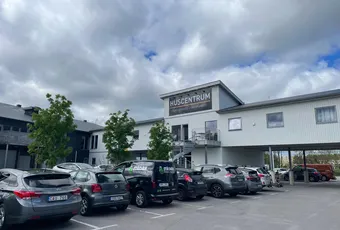 "Exterior view of Huszentrum building with parked vehicles under cloudy sky" [G]