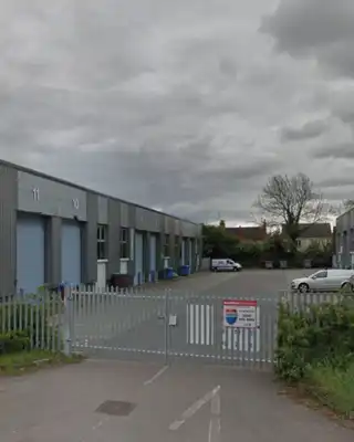 "Industrial facility with metal warehouse buildings and blue doors, surrounded by greenery under cloudy skies" [G]