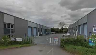 "Industrial facility with metal warehouse buildings and blue doors, surrounded by greenery under cloudy skies" [G]