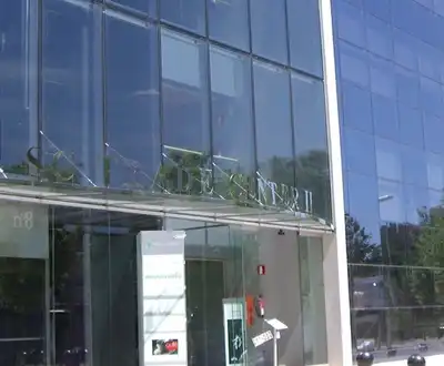 "Modern glass office building exterior with reflective facade, surrounded by trees in an urban business setting" [G]