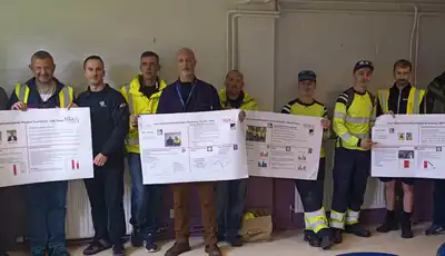 "Group of professionals in safety vests displaying informational posters in an industrial setting, representing Scott Bader's commitment to technical training." [G]