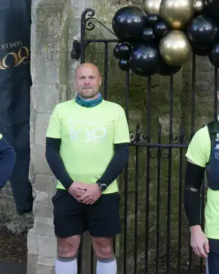 "Group of runners in bright yellow safety shirts gathered at a stone entrance with decorative balloons, representing Scott Bader community event" [G]