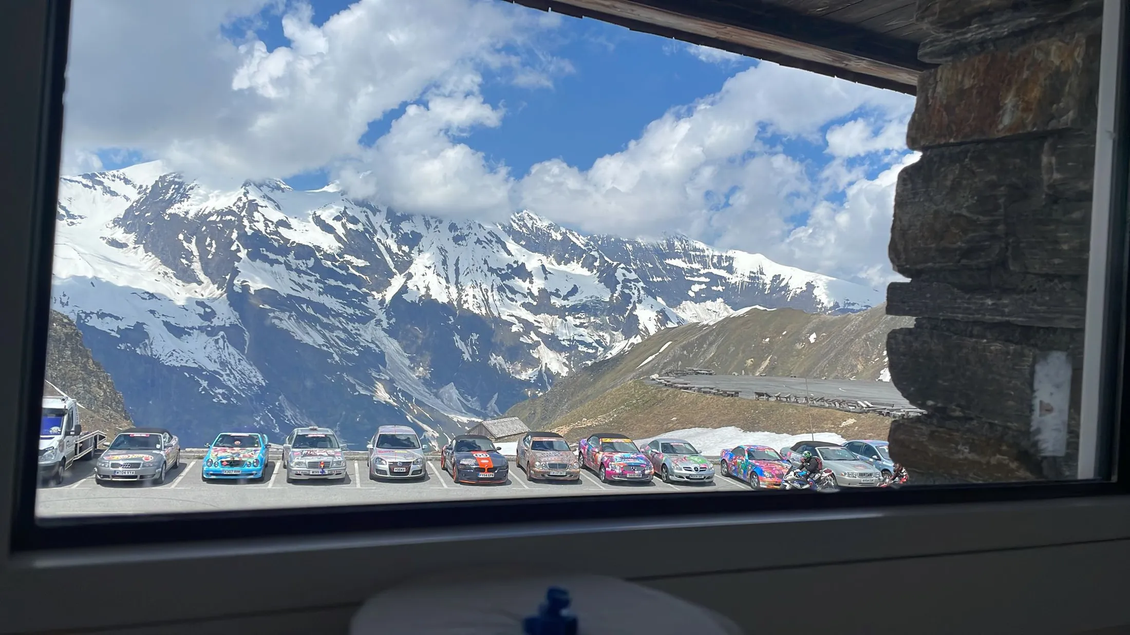 "View through window of parked cars against snow-capped mountain landscape with blue sky" [G]