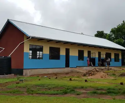 "Rural school building with blue walls and metal roof, featuring water storage tanks on the left side under cloudy sky" [G]