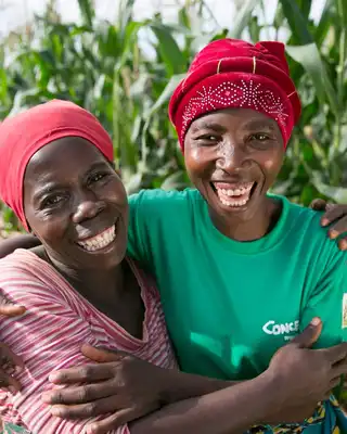 "Two agricultural workers smiling in a cornfield, standing along a dirt path with tall maize crops growing in the background." [G]