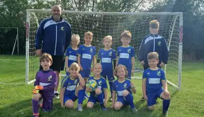 "Youth football team in blue uniforms posing on green field with soccer goal in background" [G]