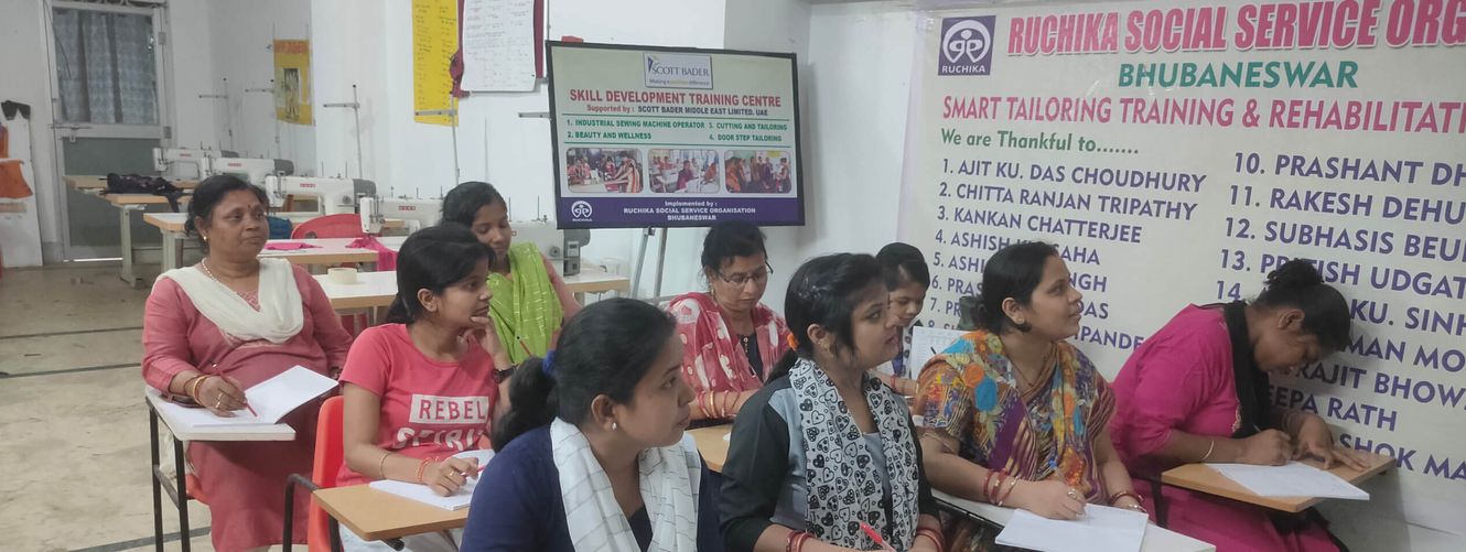 "Image of a tailoring training classroom at Ruchika Social Service Organization, with students seated at tables working on their projects." [G]
