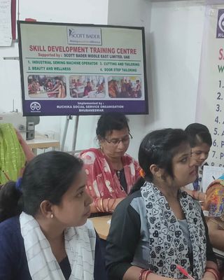 "Image of a tailoring training classroom at Ruchika Social Service Organization, with students seated at tables working on their projects." [G]