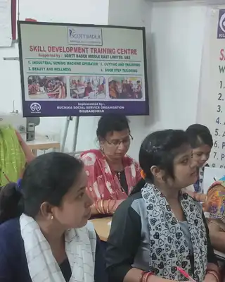 "Image of a tailoring training classroom at Ruchika Social Service Organization, with students seated at tables working on their projects." [G]