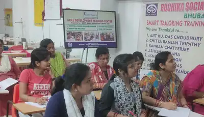 "Image of a tailoring training classroom at Ruchika Social Service Organization, with students seated at tables working on their projects." [G]