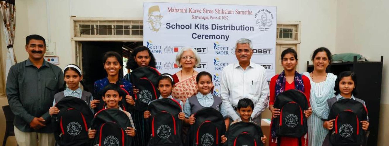 "School kits distribution ceremony with children holding backpacks, sponsored by Scott Bader and other organizations supporting education initiatives." [G]