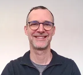 "Professional team member at Scott Bader wearing glasses and a dark zip-up jacket against a neutral background" [G]