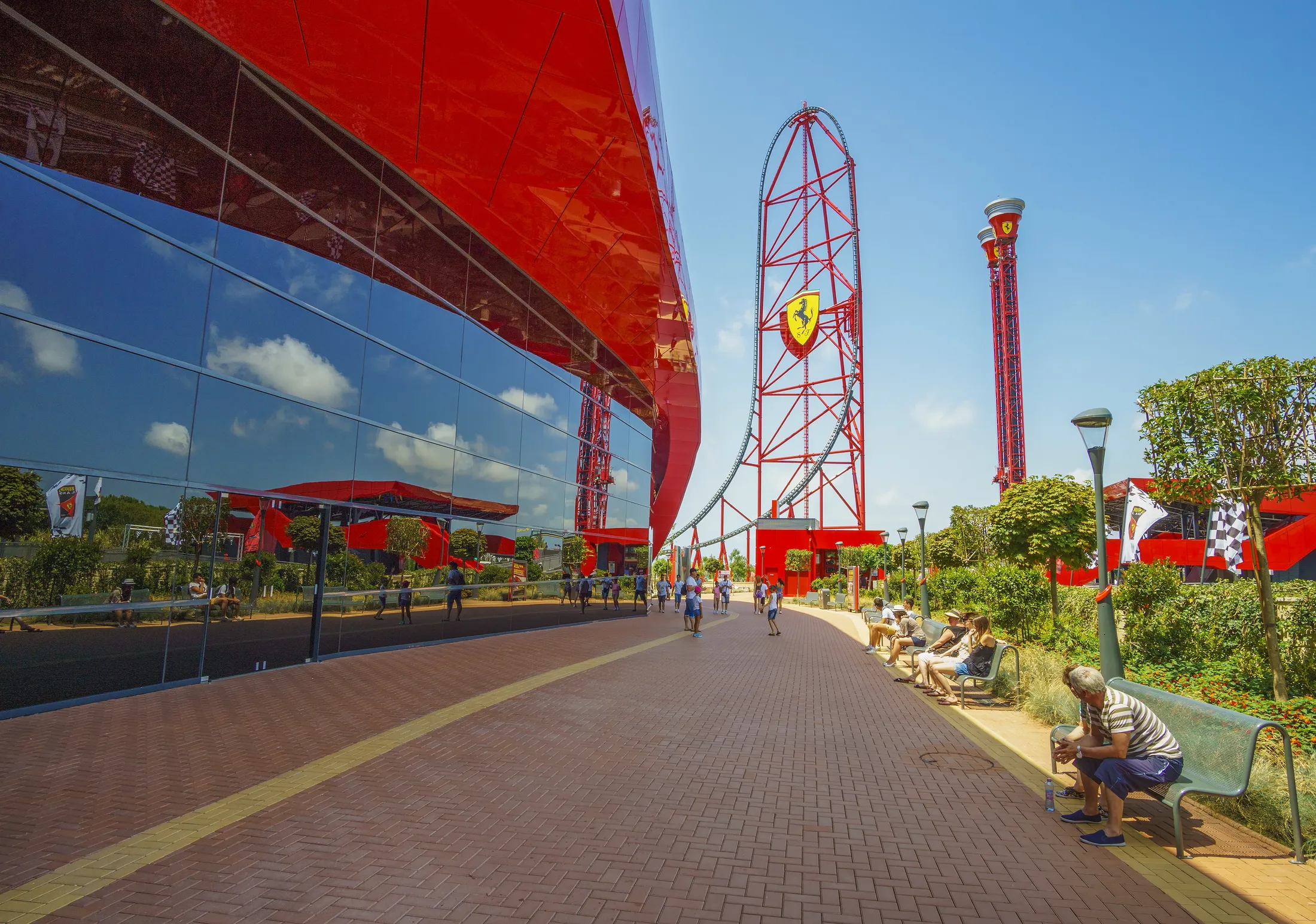 "Modern theme park with striking red roller coaster structure and glass building facade against blue sky" [G]
