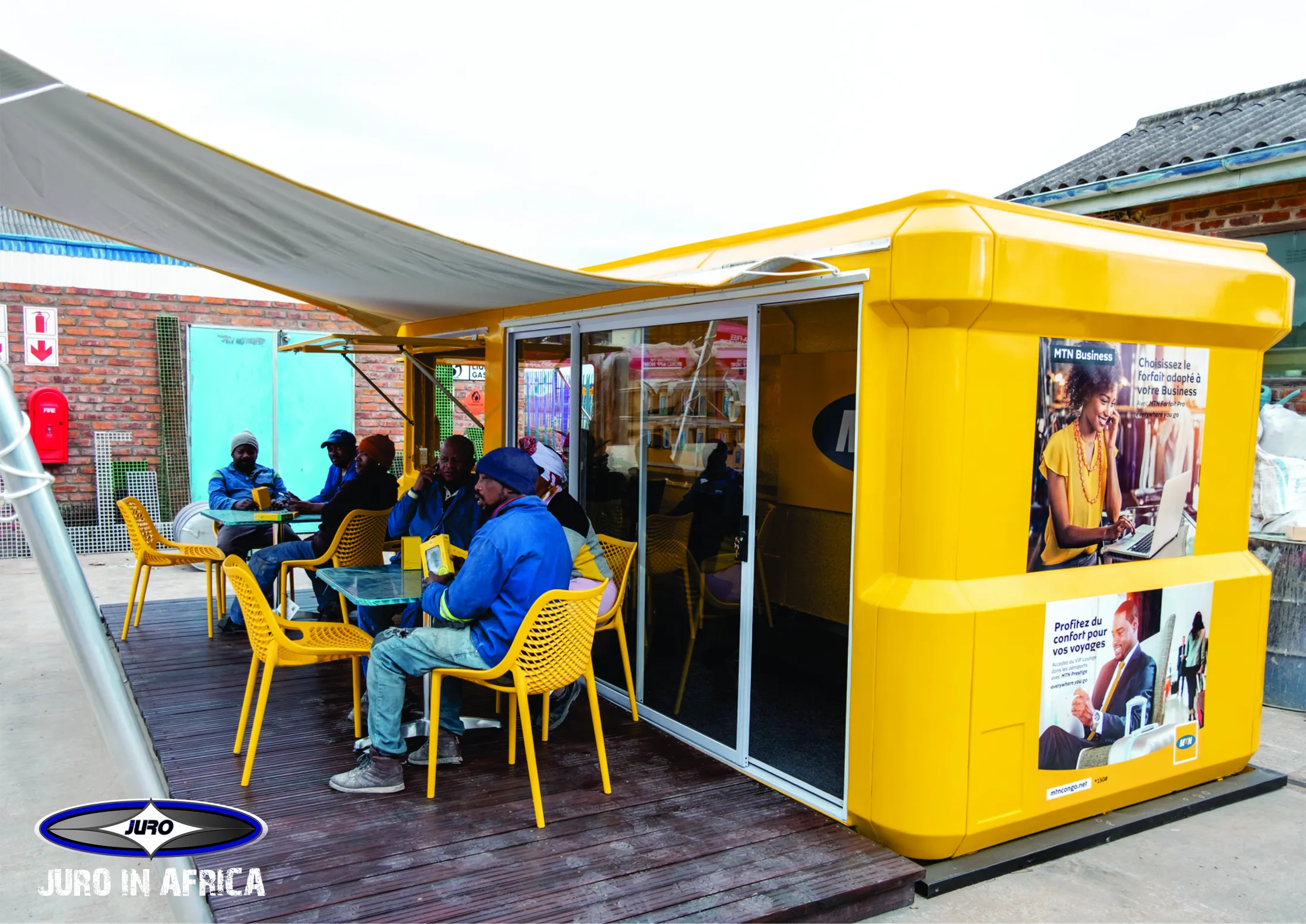 "Bright yellow community kiosk with outdoor seating area under canopy, featuring the Scott Bader logo and 'Juju in Africa' text" [G]