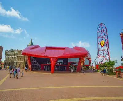 "Vibrant theme park entrance with distinctive red building and roller coaster structure against blue sky" [G]