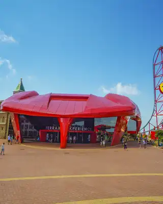 "Vibrant theme park entrance with distinctive red building and roller coaster structure against blue sky" [G]