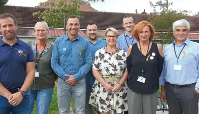"Group of professionals wearing identification badges standing outdoors in front of buildings with a grassy area" [G]