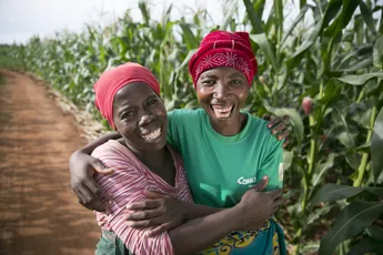 Two agricultural workers smiling in a cornfield, wearing colorful headscarves and embracing each other. [G]