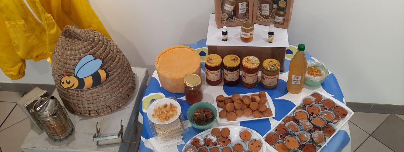 "Display of honey products and baked goods on a blue tablecloth with decorative beehive and Scott Bader packaging visible in wooden display case" [G]