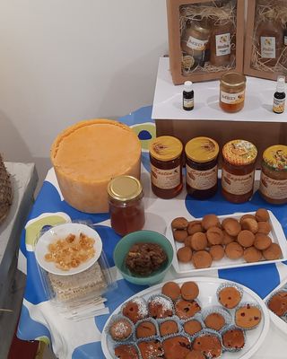 "Display of honey products and baked goods on a blue tablecloth with decorative beehive and Scott Bader packaging visible in wooden display case" [G]