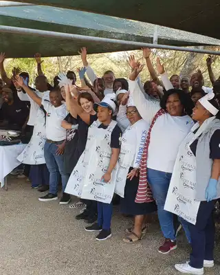 "Group of enthusiastic team members at outdoor event with hands raised, some wearing aprons and Scott Bader branded shirts." [G]